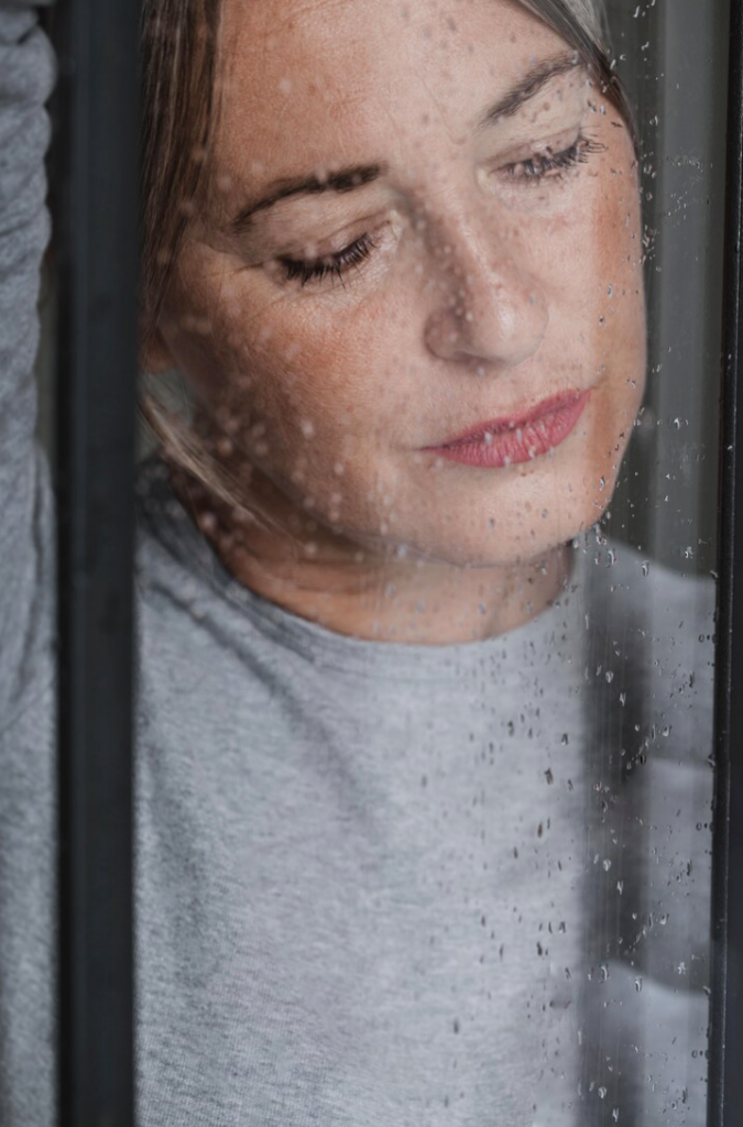 white woman riding waves of grief looking out a window with rain droplets