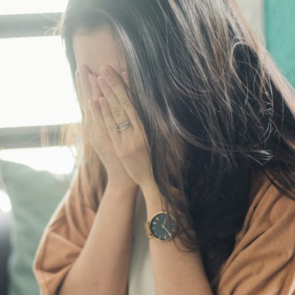 Woman hiding her face with both hands, wearing a watch and rings, seated by a window.